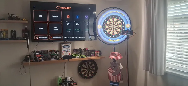 A home darts corner with DartsKEY on a wall TV and a phone mounted on the dartboard's light-ring stand