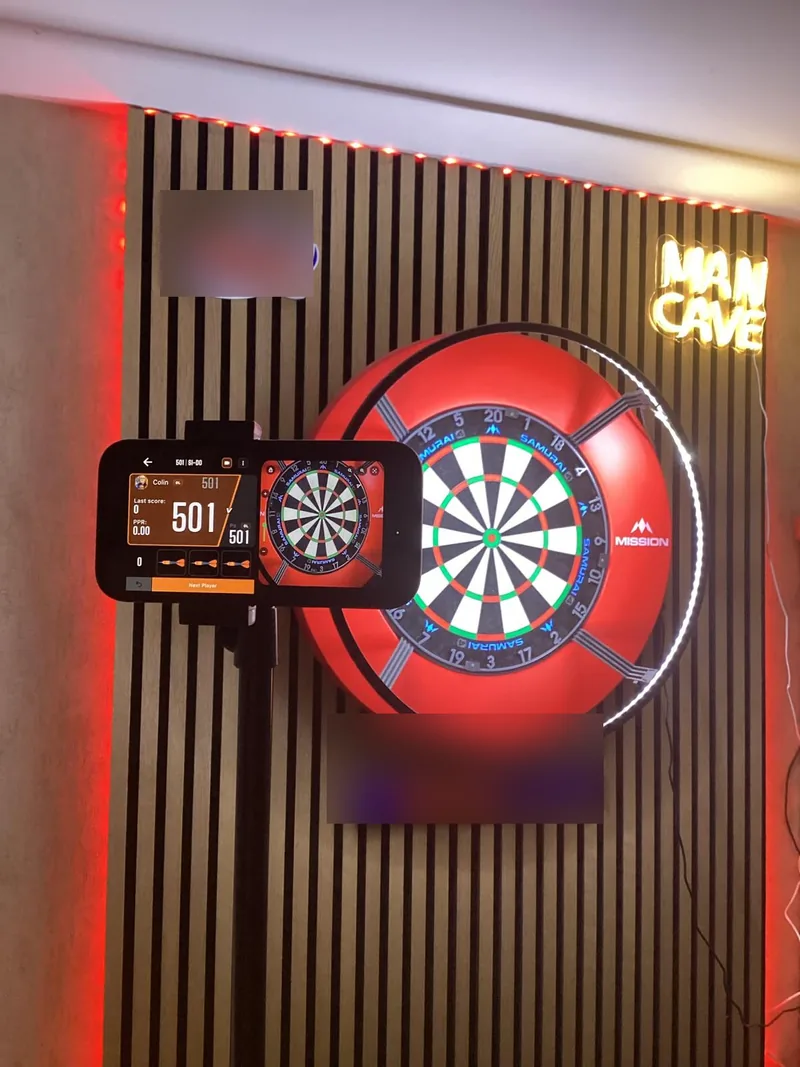 A red-lit dartboard in a man cave with a phone on a stand beside it showing a DartsKEY 501 game