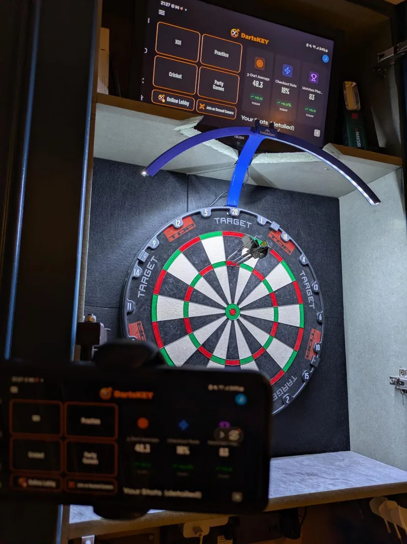 A dartboard with a tablet above it and a phone mount in front, both showing DartsKEY