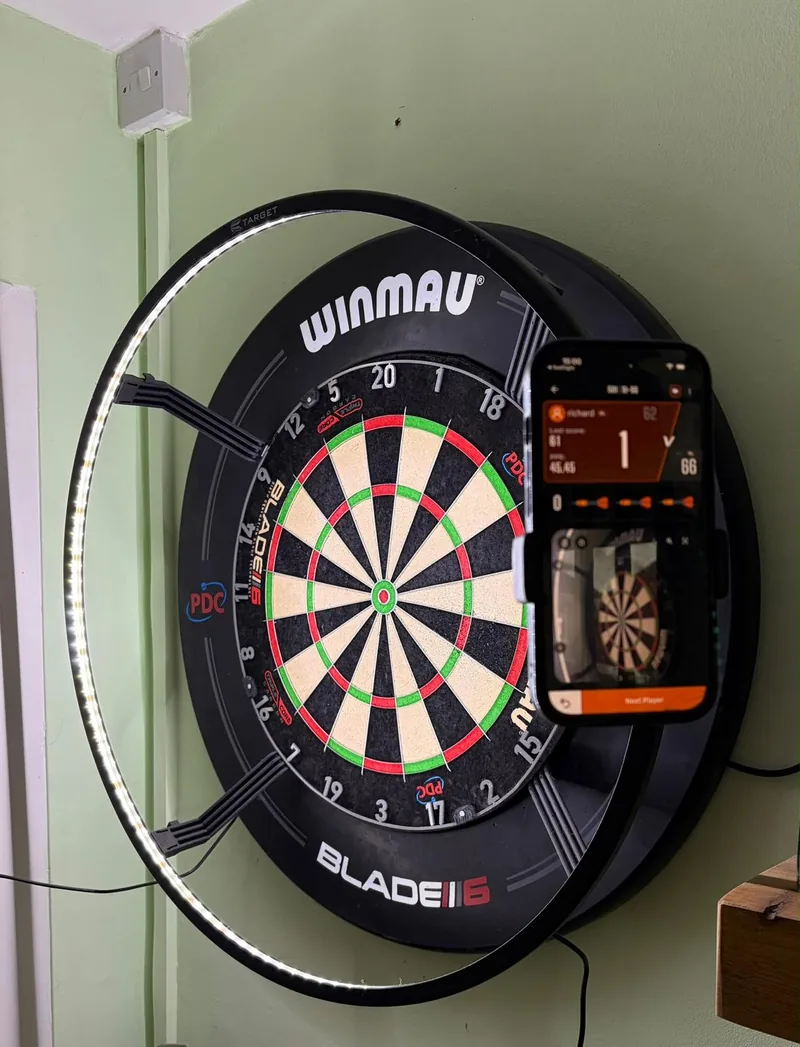 A phone clamped to a dartboard surround, live-scoring a DartsKEY game