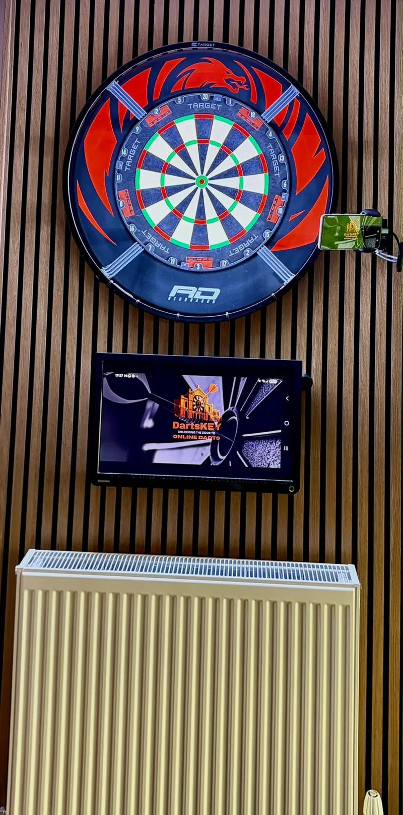 A dartboard on a slatted wall with a tablet below it showing DartsKEY